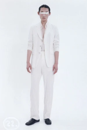 An adult Asian man in a white suit stands in front of a white backdrop wearing black shoes, shot with studio lighting. Minimalist fashion.