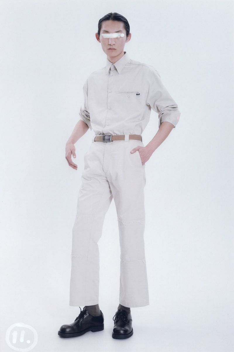A stylish adult male poses in a photo studio wearing a beige button-down shirt, matching trousers, black shoes, and a white band across his eyes.