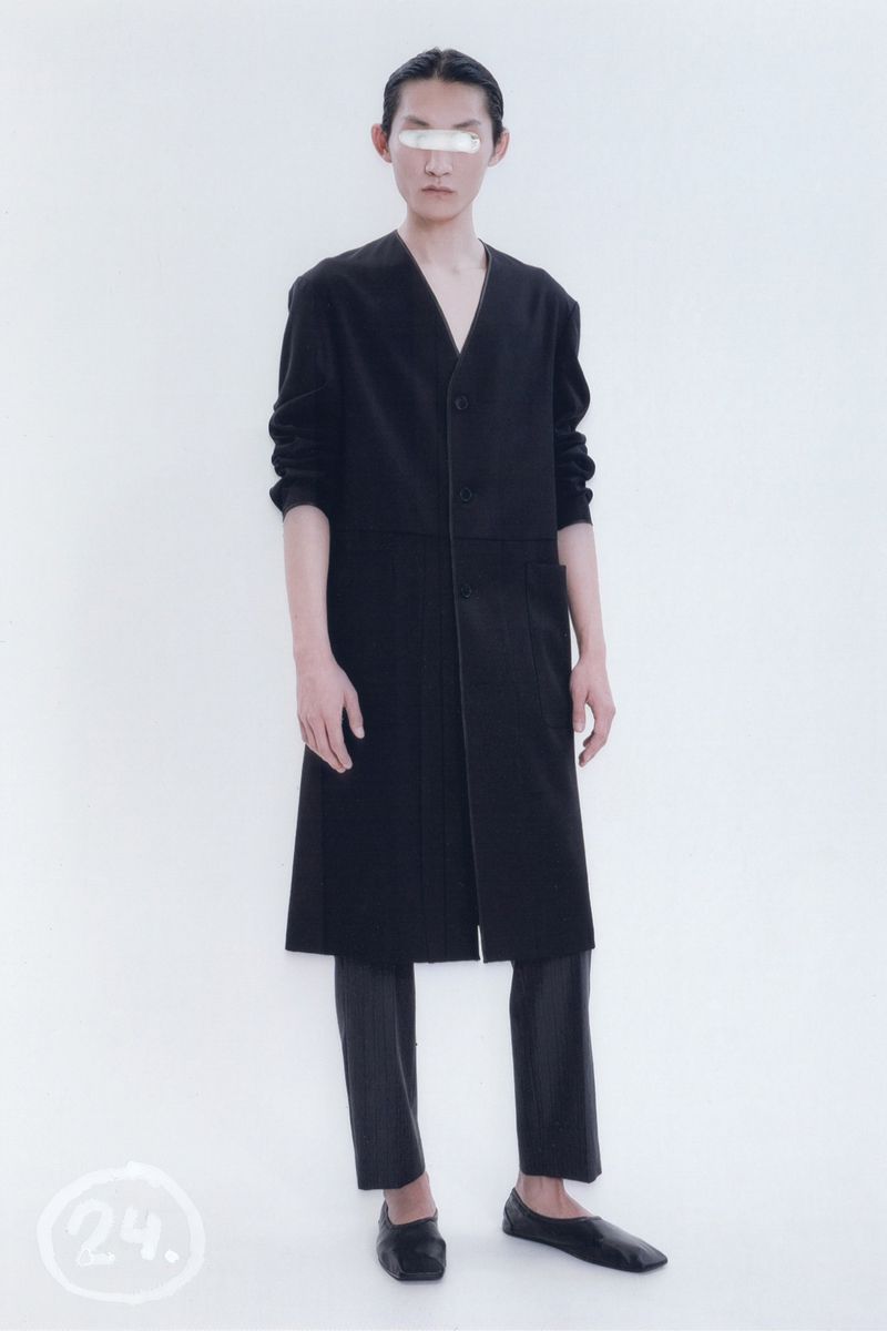 Person in black coat and pants with facial covering posing in a bright photo studio setting. Fashion editorial photograph.