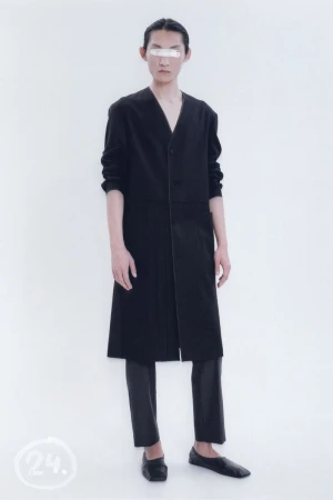 Person in black coat and pants with facial covering posing in a bright photo studio setting. Fashion editorial photograph.