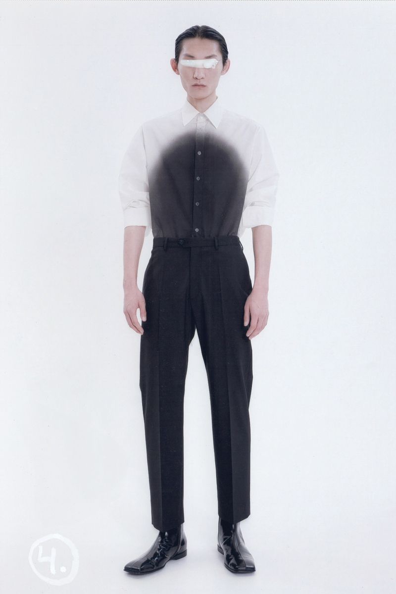 Asian male model poses in white shirt, black trousers, and patent boots against a light backdrop, fashion forward clothing and eyewear.