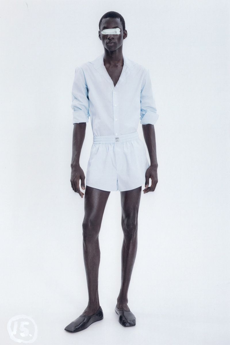 An adult male stands in a photo studio, wearing a light-blue shirt, light-blue shorts, and black loafers against a bright-white background.