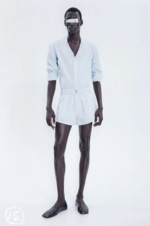 An adult male stands in a photo studio, wearing a light-blue shirt, light-blue shorts, and black loafers against a bright-white background.