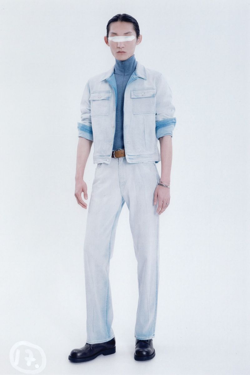 Fashion model in denim jacket and pants stands in a photo studio. The model has a neutral expression with a strip across the eyes.
