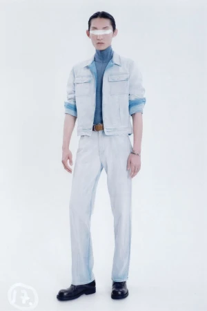 Fashion model in denim jacket and pants stands in a photo studio. The model has a neutral expression with a strip across the eyes.