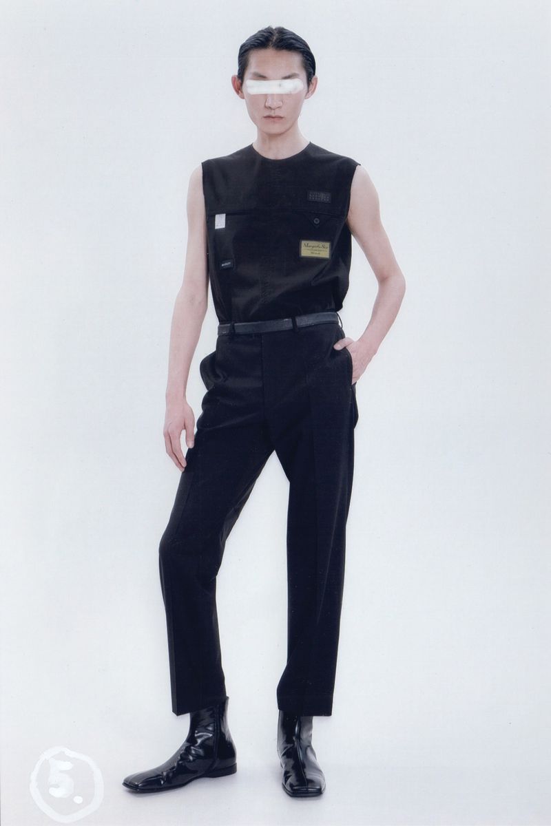 An Asian male fashion model in a black vest and trousers with a white censor bar on his eyes, posing in a bright white photo studio.