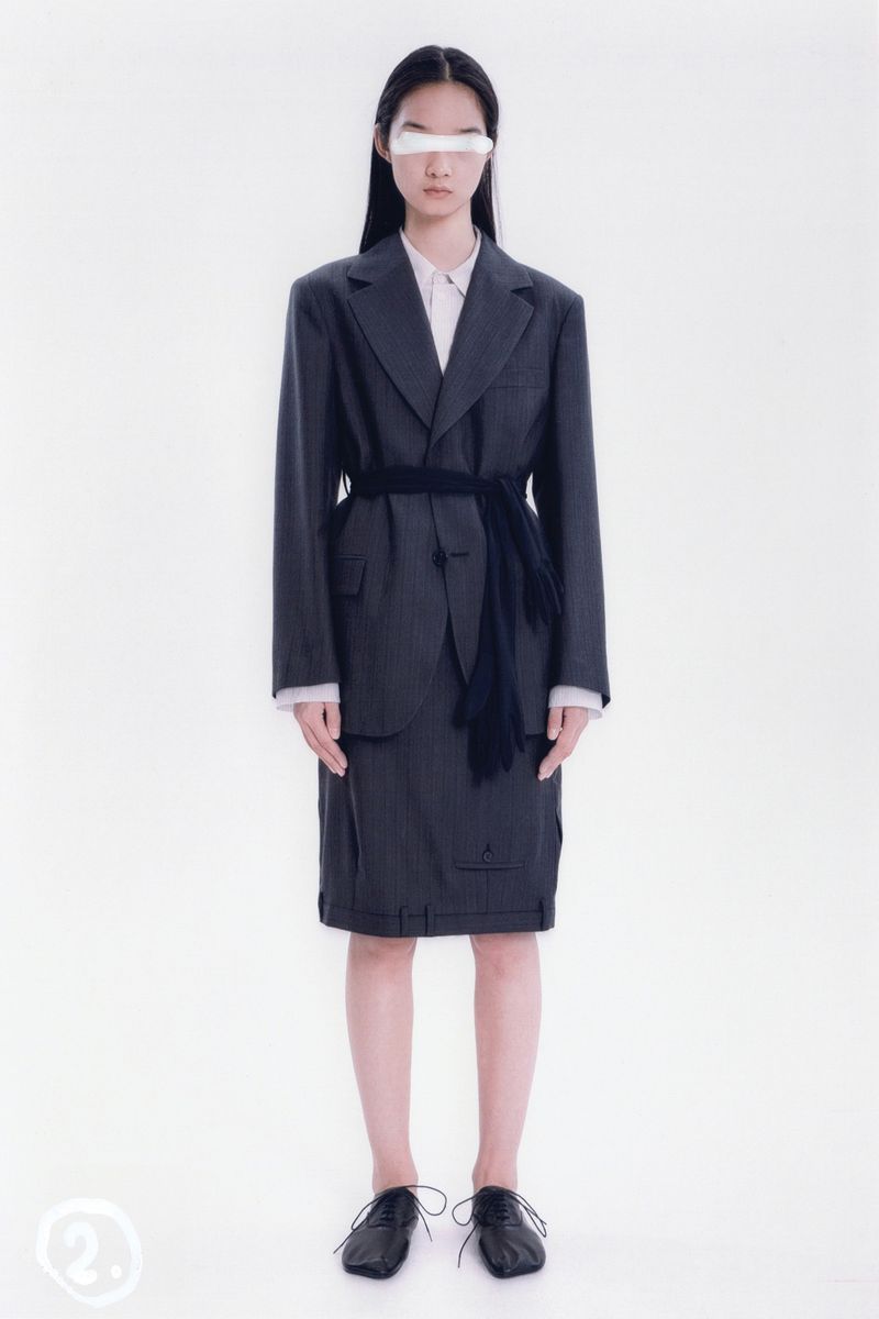 An Asian female model poses in a dark pinstripe skirt suit, white shirt, dark shoes, and a white covering over her eyes in a photo studio.