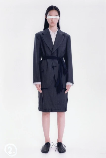An Asian female model poses in a dark pinstripe skirt suit, white shirt, dark shoes, and a white covering over her eyes in a photo studio.