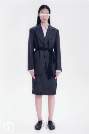 An Asian female model poses in a dark pinstripe skirt suit, white shirt, dark shoes, and a white covering over her eyes in a photo studio.