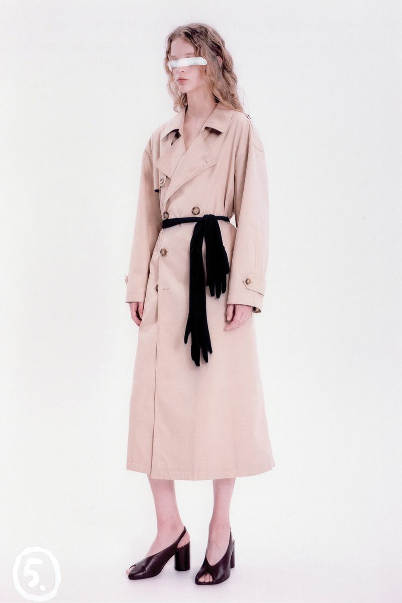 A stylish woman in a tan trench coat and black gloves poses in a studio with bright lighting, creating a high-fashion editorial shot.