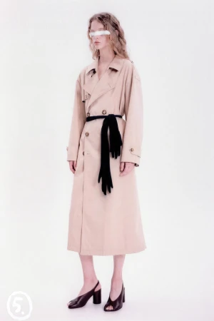 A stylish woman in a tan trench coat and black gloves poses in a studio with bright lighting, creating a high-fashion editorial shot.