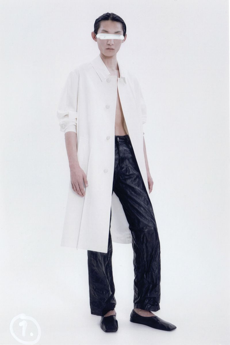 Full-length studio shot of a person in a white coat and dark pants, with a white bar obscuring the eyes against a clean white background.