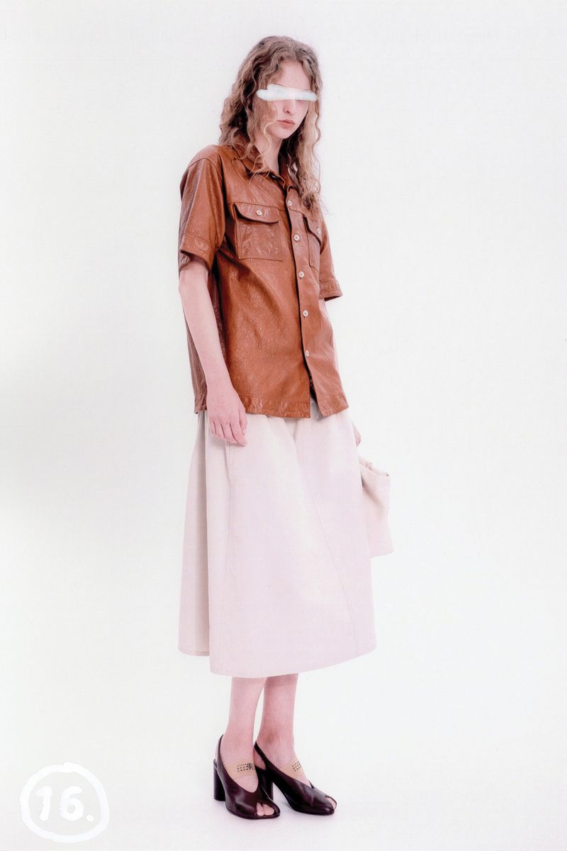 A model poses in a studio with soft, diffused light wearing a brown leather shirt, beige skirt, and chunky black heels with metallic details.