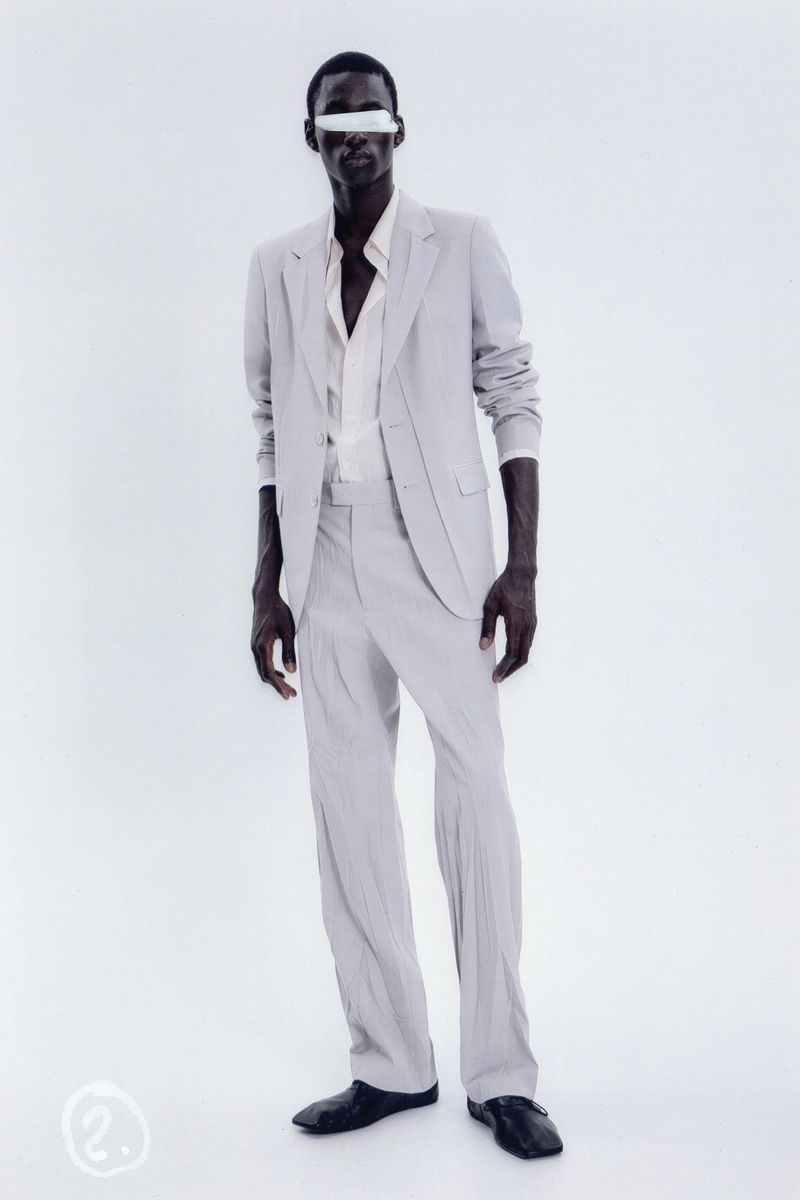 An African American adult male stands in a light grey suit with a light shirt and dark shoes against a light backdrop in a studio shot.