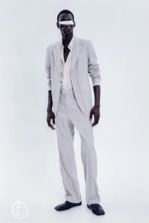 An African American adult male stands in a light grey suit with a light shirt and dark shoes against a light backdrop in a studio shot.