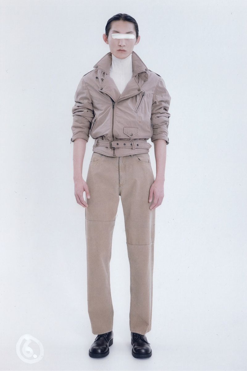 Full-length studio shot of a slender person with white band across the eyes, wearing a khaki jacket and pants, on a white backdrop.