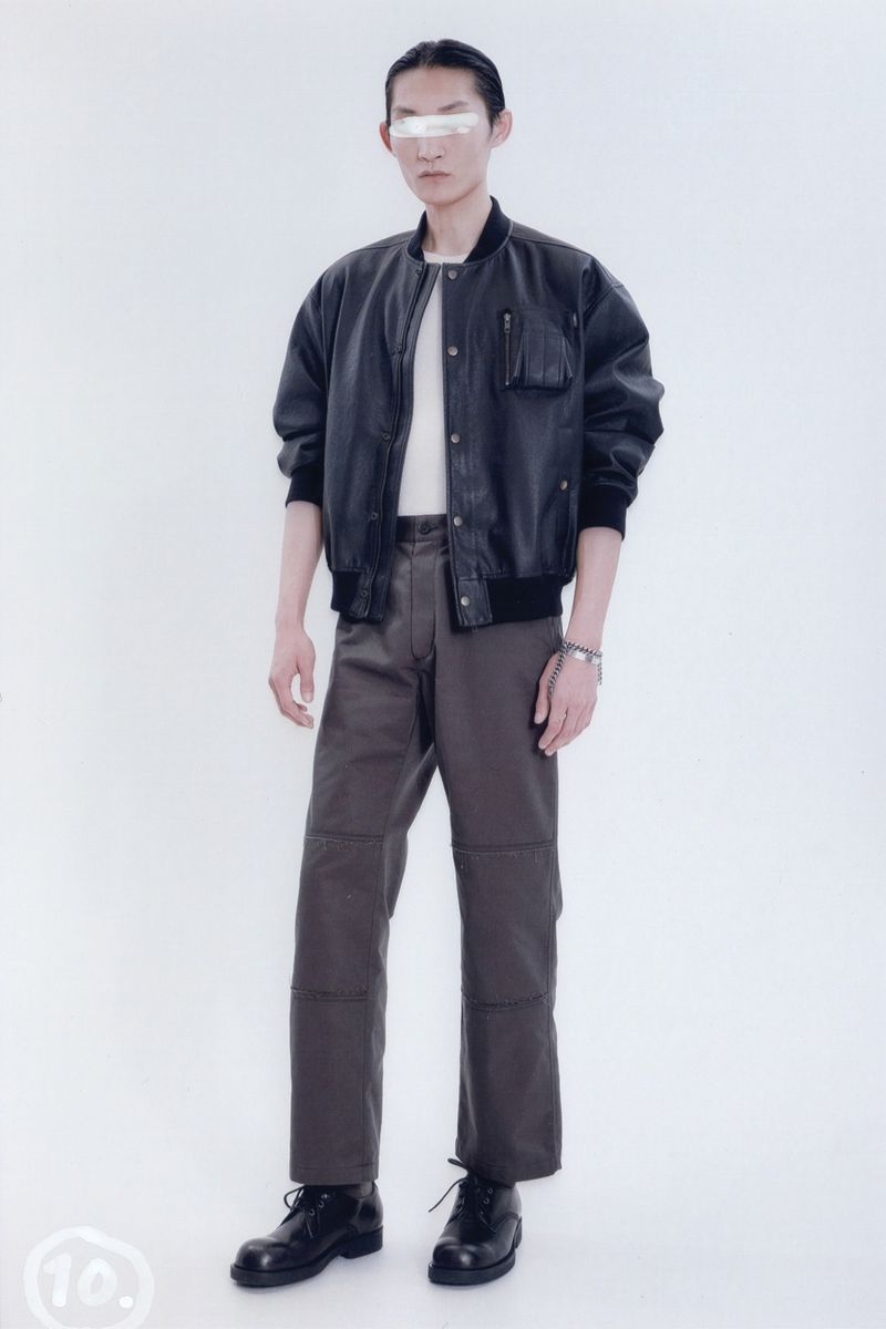 Full-length studio shot of a young adult male fashion model wearing a black bomber jacket, grey pants, and black boots on a white backdrop.