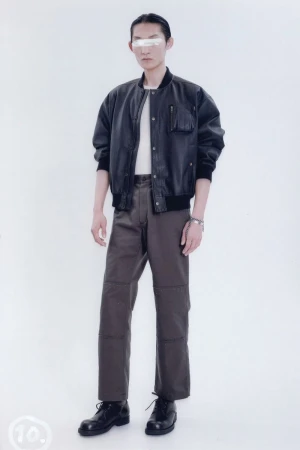 Full-length studio shot of a young adult male fashion model wearing a black bomber jacket, grey pants, and black boots on a white backdrop.