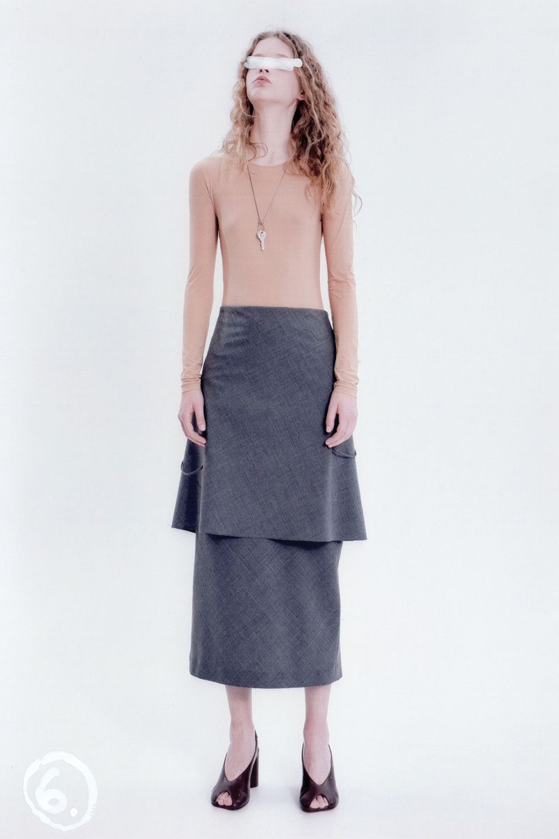 Fashion shot of a woman with curly hair wearing a long-sleeve top and a layered midi skirt, accessorized with heels and a necklace, against a white backdrop.