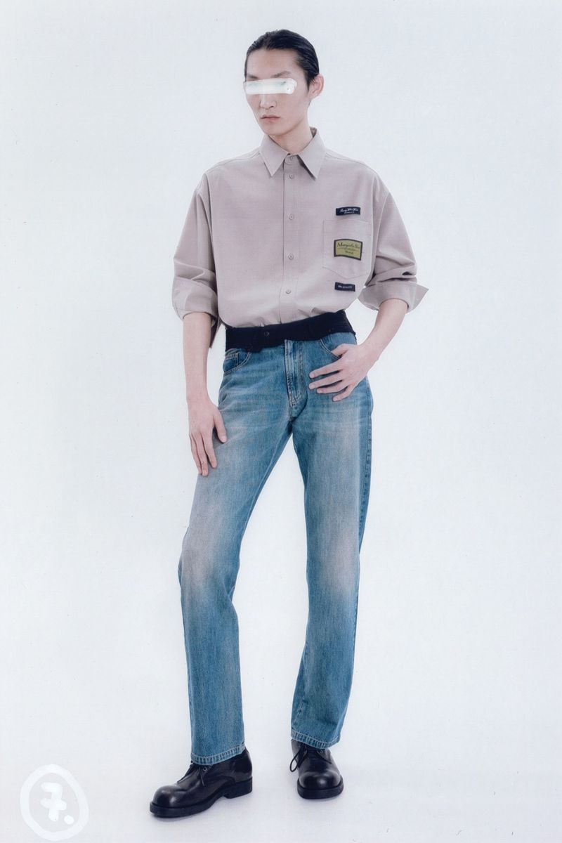 Adult male fashion model poses in a studio wearing a beige button-down shirt, jeans, and black shoes, showcasing a minimalist fashion style.