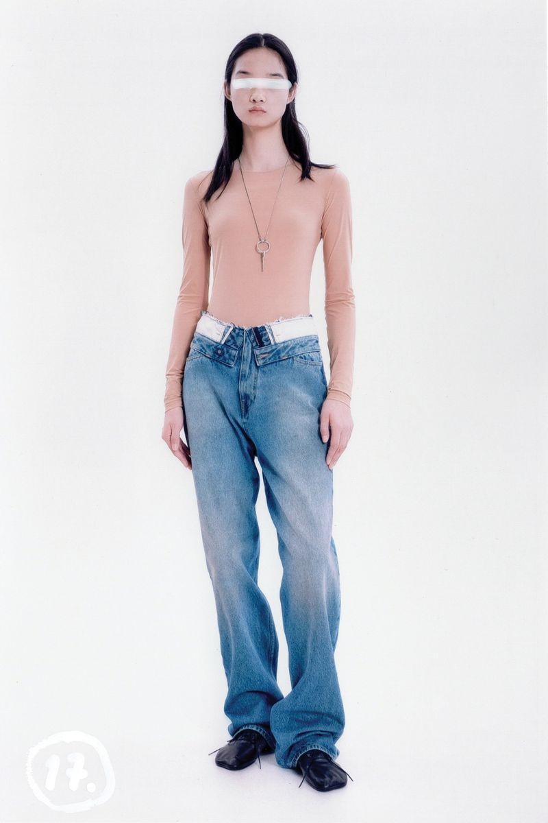 A young adult poses in a studio with a light skin tone, wearing a tan top, light blue jeans, and black shoes against a white backdrop.
