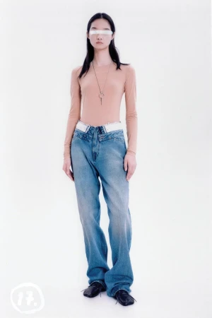 A young adult poses in a studio with a light skin tone, wearing a tan top, light blue jeans, and black shoes against a white backdrop.