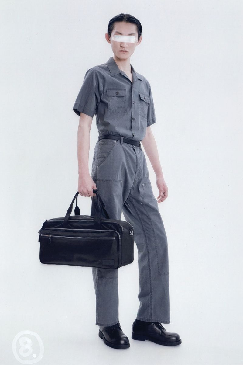 Studio shot of a person in a grey short-sleeved shirt and grey pants, holding a black bag and wearing black shoes.