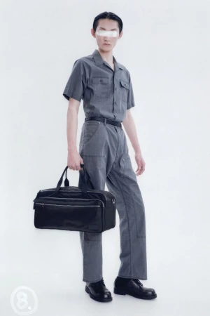 Studio shot of a person in a grey short-sleeved shirt and grey pants, holding a black bag and wearing black shoes.