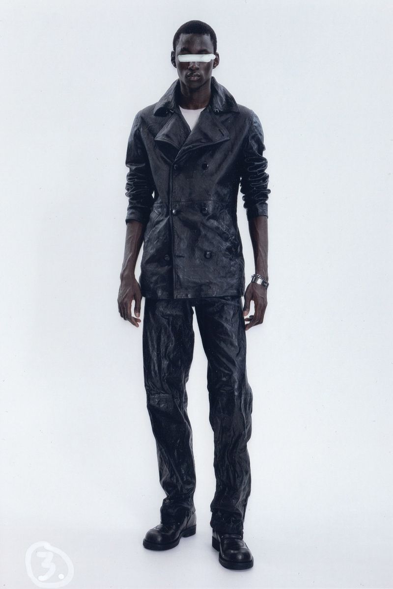 Dark-skinned male fashion model in black leather jacket and pants with white block over eyes, standing in a bright white photo studio.