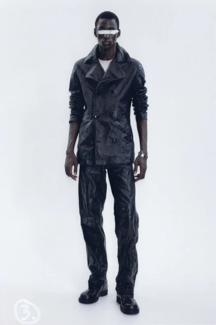 Dark-skinned male fashion model in black leather jacket and pants with white block over eyes, standing in a bright white photo studio.