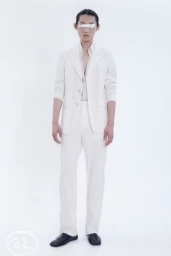 Model wearing an all-white tailored suit and black shoes with white paint over his eyes against a bright white studio backdrop.
