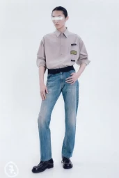 Model wearing a beige collared shirt, washed blue jeans, and black leather shoes against a plain white studio background.