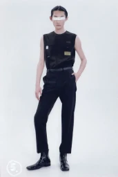 Model wearing a black sleeveless top and black trousers with a white band covering their eyes against a seamless white background.