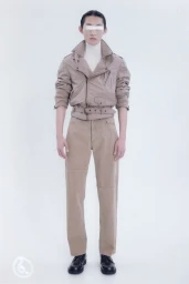 Model wearing a beige belted biker jacket and matching trousers with a white eye covering against a white seamless backdrop.