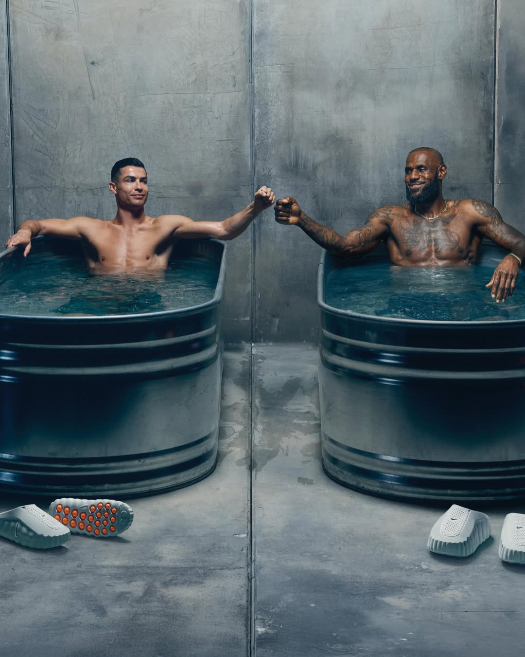 Cristiano Ronaldo and LeBron James sitting in metal ice bath tubs and fist-bumping in a concrete room.