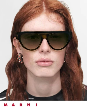 Close-up portrait of model Laura Rudd with red hair wearing oversized dark flat-top sunglasses against a white background.