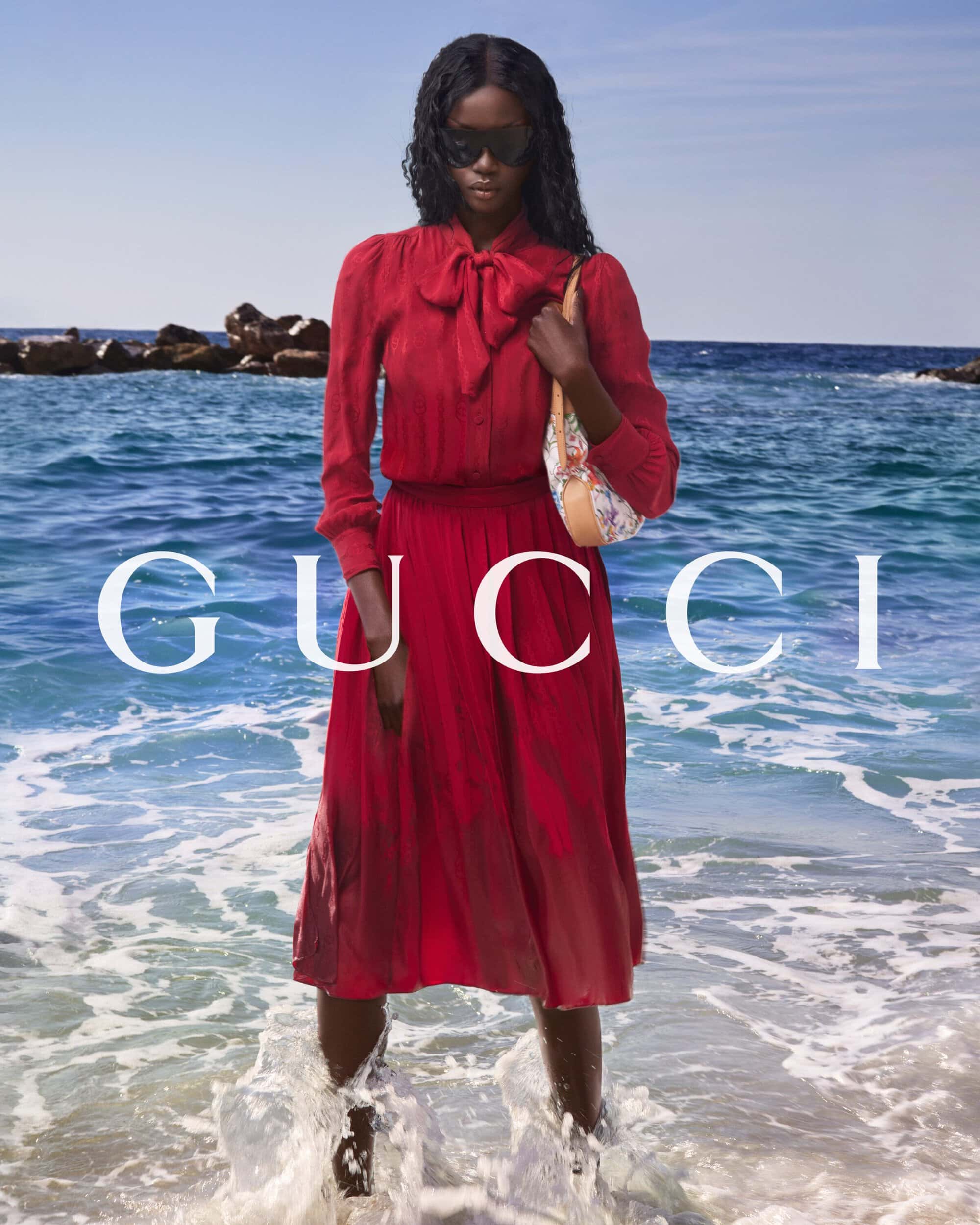 Model Anok Yai standing in ocean surf wearing a red long-sleeve bow dress and sunglasses for the Gucci Summer 2026 campaign.