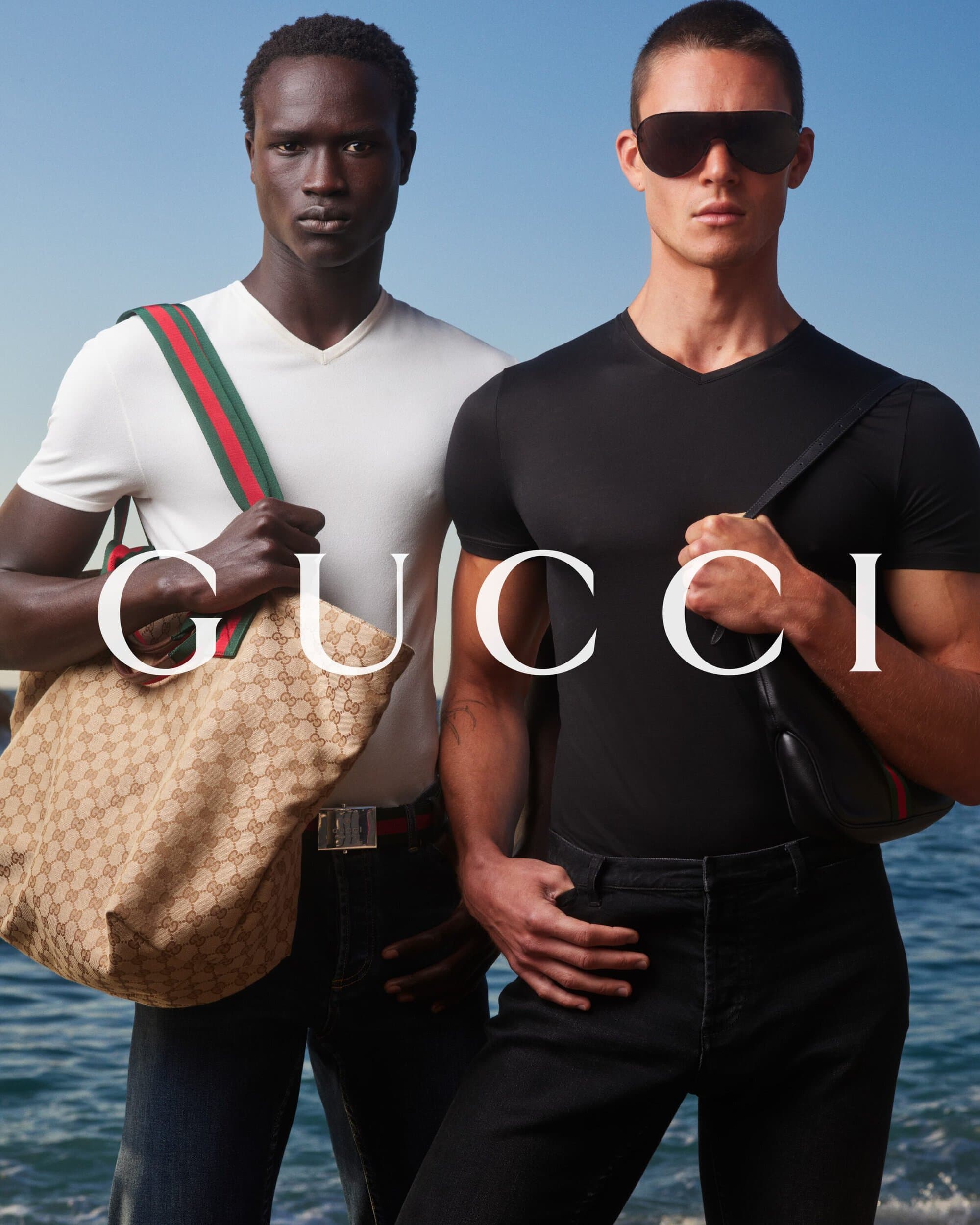 Two male models posing against an ocean backdrop, one holding a canvas tote bag and wearing a white t-shirt, the other wearing sunglasses and black clothing.
