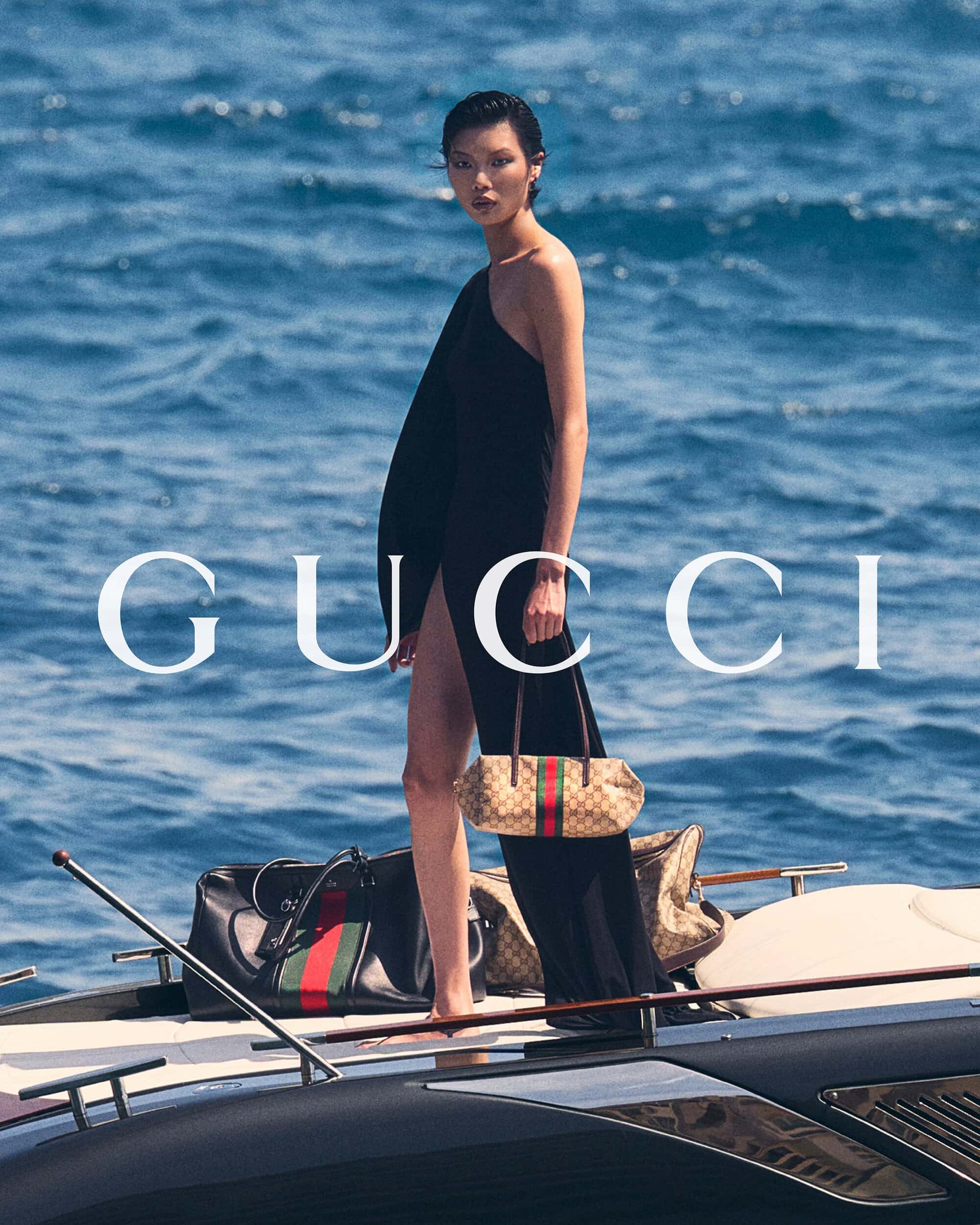 A model standing on a boat deck wearing a black one-shoulder high-slit dress and holding a Gucci monogram handbag against blue open water.