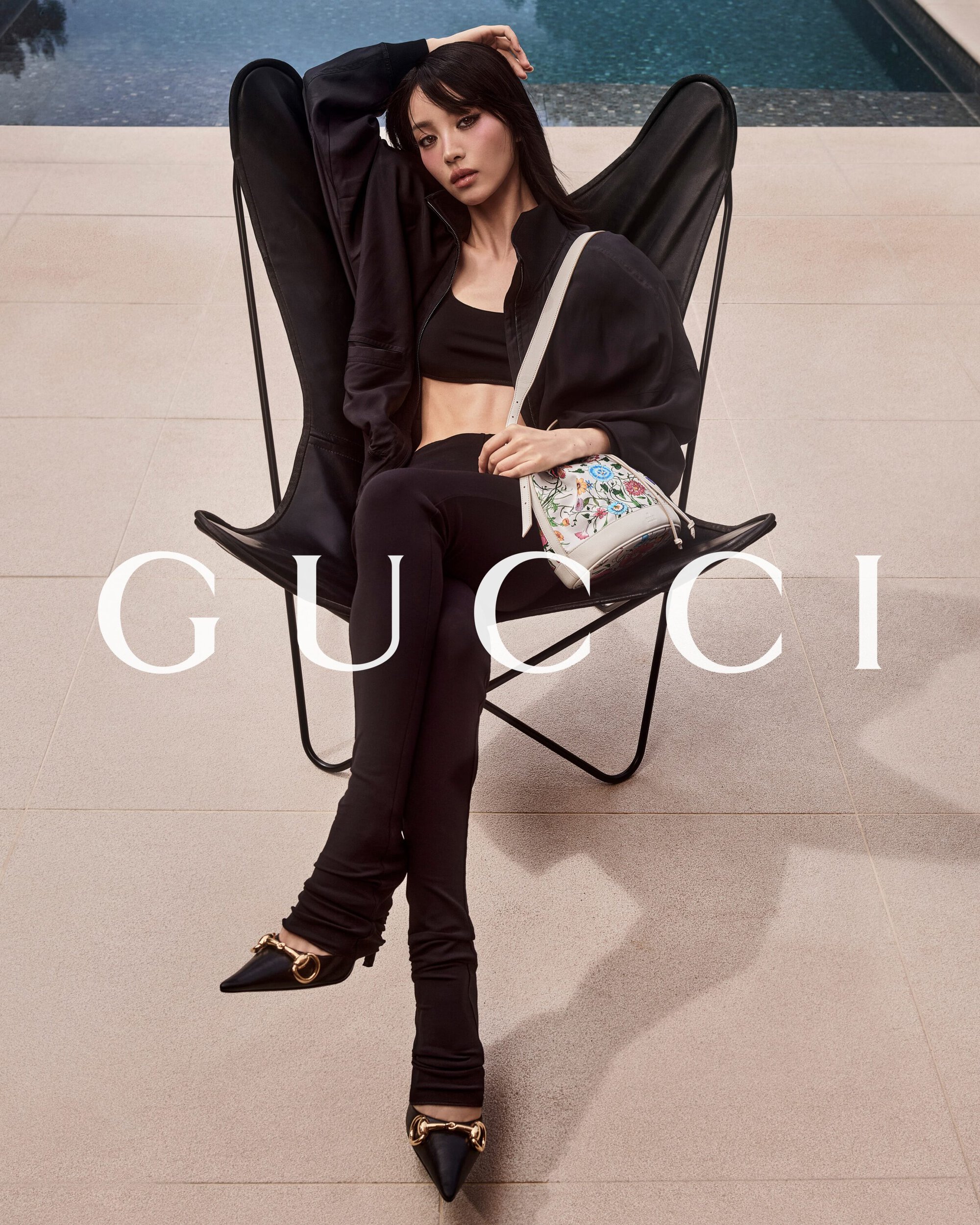 Model Tian Xi Wei seated in a black butterfly chair by a pool wearing black attire and holding a white patterned bucket bag for Gucci.