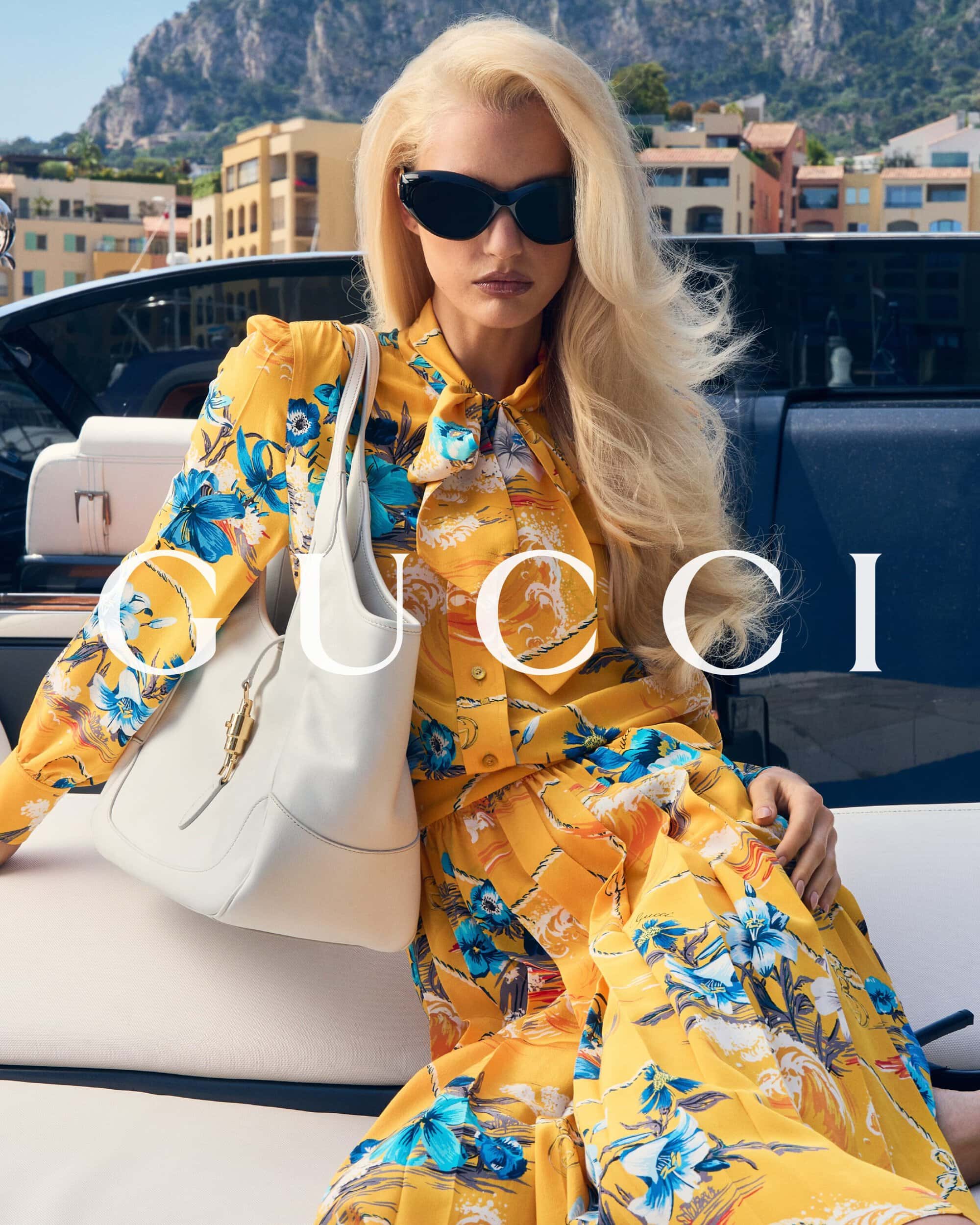 Model Amelia Gray wearing a yellow printed floral dress and black sunglasses with a white leather shoulder bag on a yacht.