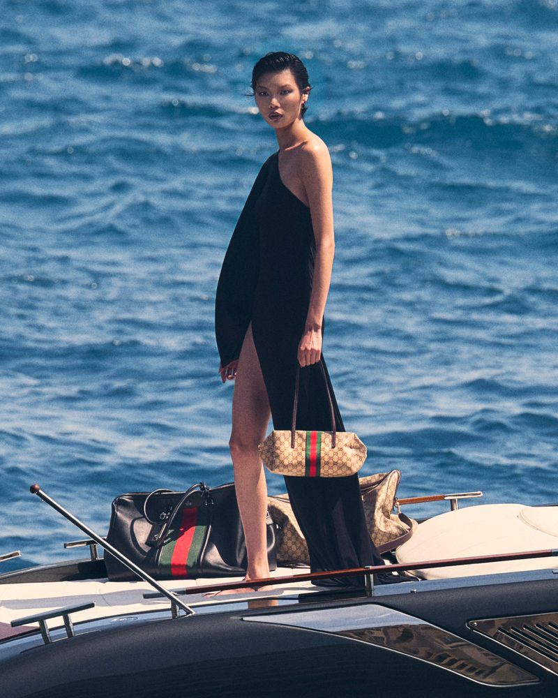 Model posing in a black one-shoulder slit gown holding a monogram handbag on a boat deck overlooking deep blue ocean water.