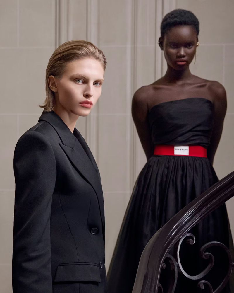 Two fashion models posing near a staircase railing in tailored black garments with a bright red waist belt accent.