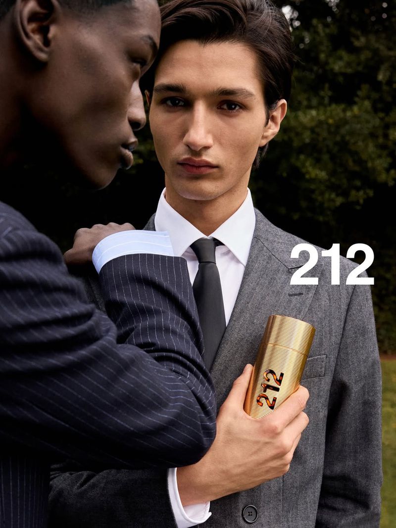 A man in a grey suit and black tie holds a gold 212 perfume bottle next to a man in a navy pinstripe suit outdoors.
