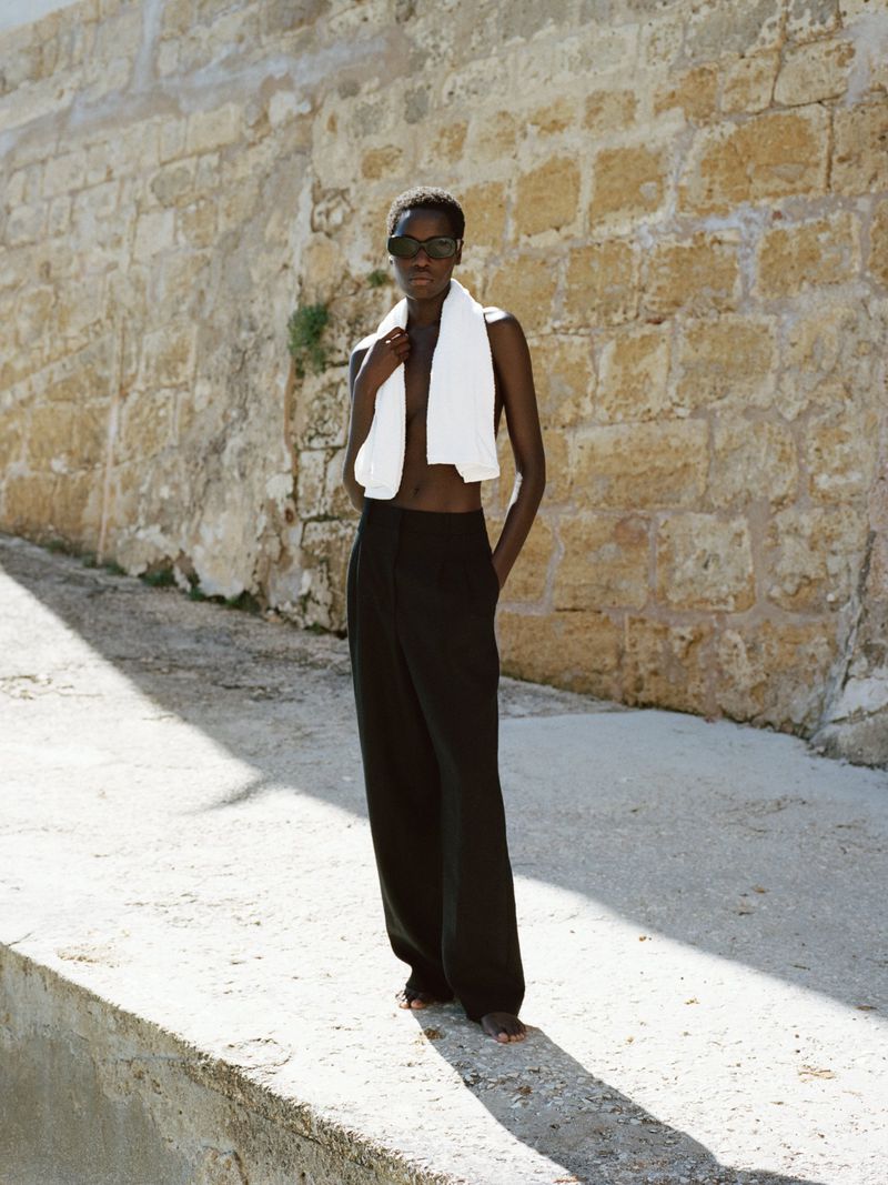 Model Awar Odhiang wearing black wide-leg trousers, sunglasses, and a white towel around her neck against a sunlit stone wall.
