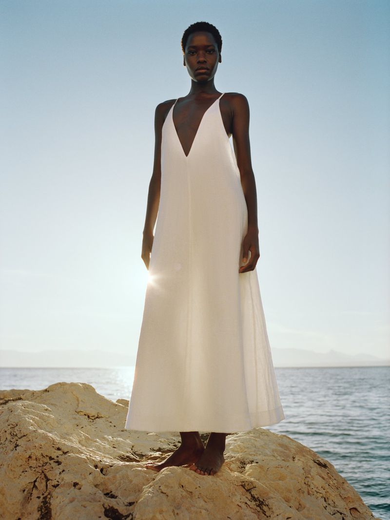 Model Awar Odhiang stands barefoot on coastal rocks overlooking the sea wearing a white plunging V-neck sleeveless maxi dress.