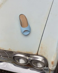A light blue open-toe mule shoe resting on the faded hood of a vintage car with rust patina and chrome round headlights.