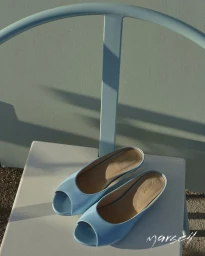Pair of light blue open-toe mule shoes resting on a pale mint metal chair in bright sunlight with crisp shadows.