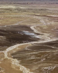 High-angle view of a meandering dry creek bed winding across an arid desert landscape with layered earth tones.