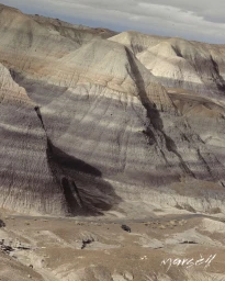 Stratified desert hills with visible purple and beige sediment layers under a soft sky in a vast arid landscape.